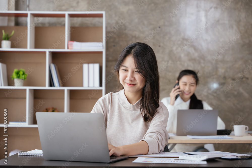 Young business women working on laptop with happy and smile face with co-worker is discussing business by smartphone behind her in office spec. Young business women working on laptop with happy and smile face with co-worker is discussing business by smartphone behind her in office spec.