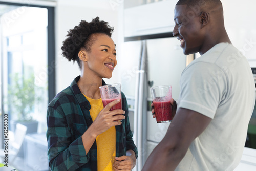 Happy african american mid adult couple holding smoothie in glasses looking at each other in kitchen