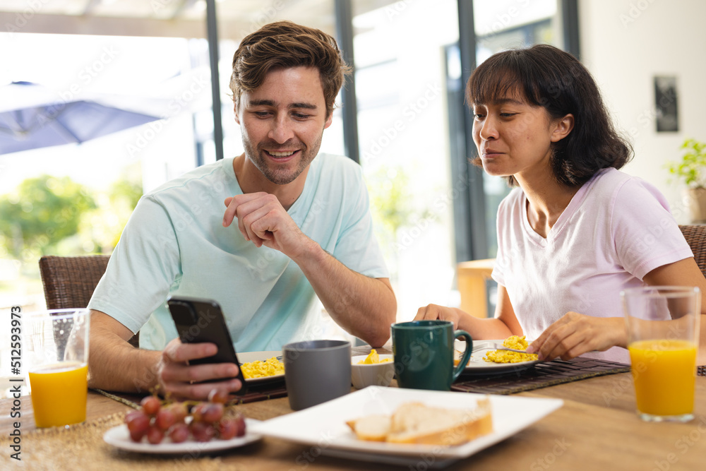 Caucasian boyfriend showing mobile phone to asian girlfriend while having breakfast on dining table Caucasian boyfriend showing mobile phone to asian girlfriend while having breakfast on dining table
