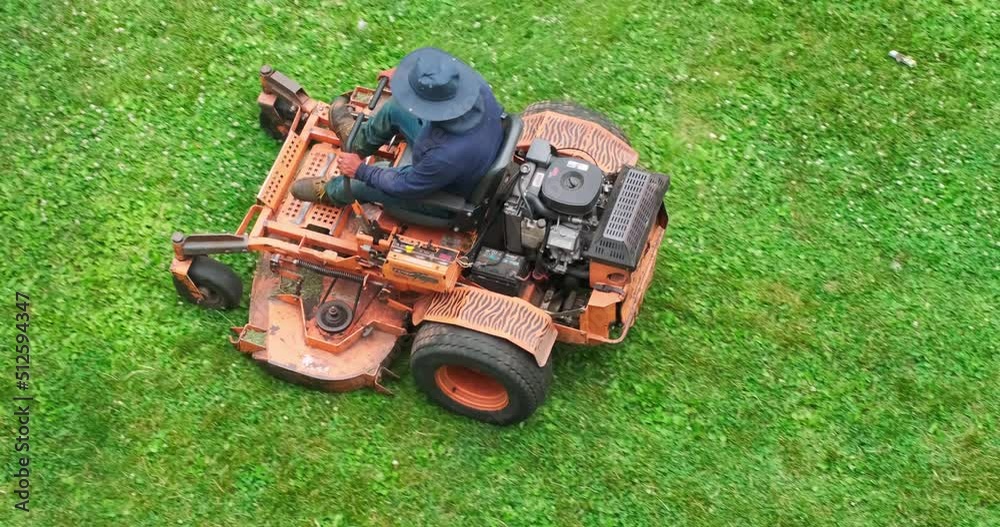 Top down shot a landscaper on an orange commercial riding lawn mower cutting grass on a cloudy summer afternoon.