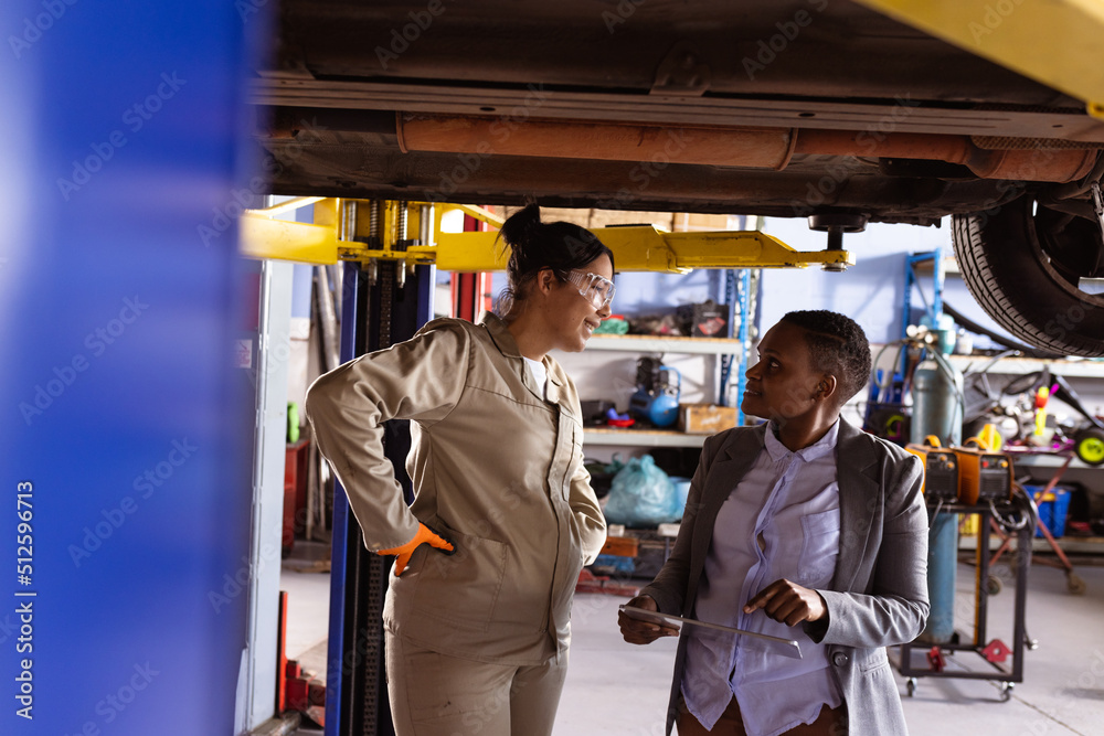 Multiracial mid adult female engineers discussing work under car lift in workshop, copy space Multiracial mid adult female engineers discussing work under car lift in workshop, copy space