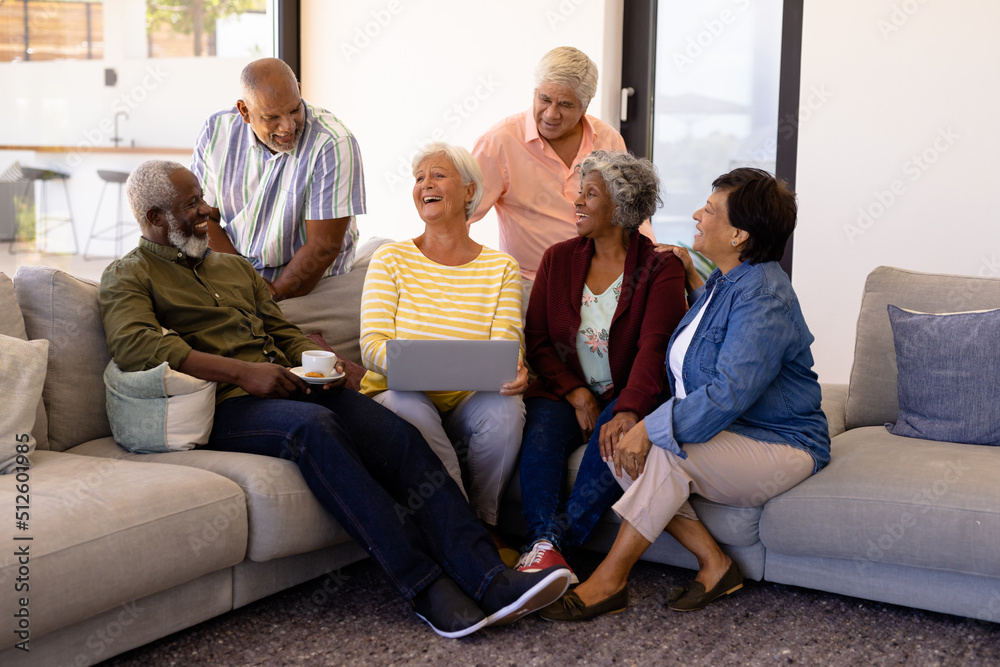 Multiracial happy senior woman with laptop sitting with friends on sofa in nursing home