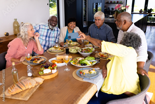 Multiracial senior woman showing smart phone to friends while having lunch at dining table