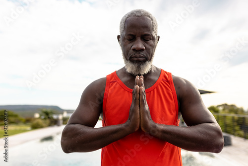African american senior man with eyes closed meditating in prayer position against sky in yard