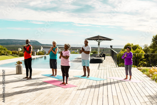 Multiracial seniors meditating while standing on hardwood floor at poolside against sky in summer