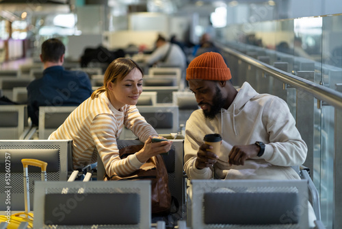 Beautiful European woman asks handsome African American man to help connect wifi. Young couple watching video or photo in social media on mobile phone together. New acquaintances during flight delay.