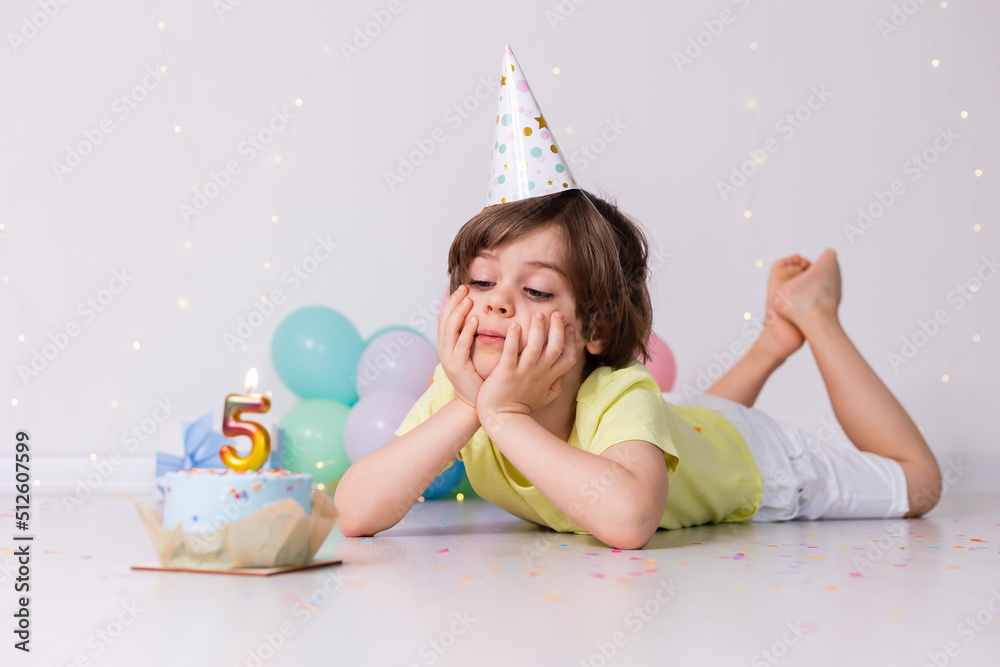 Fototapeta premium Cute little birthday boy in hat, cake with candle five, balloons, happy birthday
