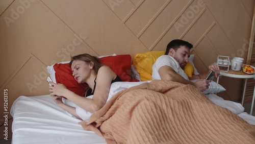 A married couple is lying in bed with their faces turned away from each other using phones. Family quarrel, touchy spouses lie separately after swearing photo