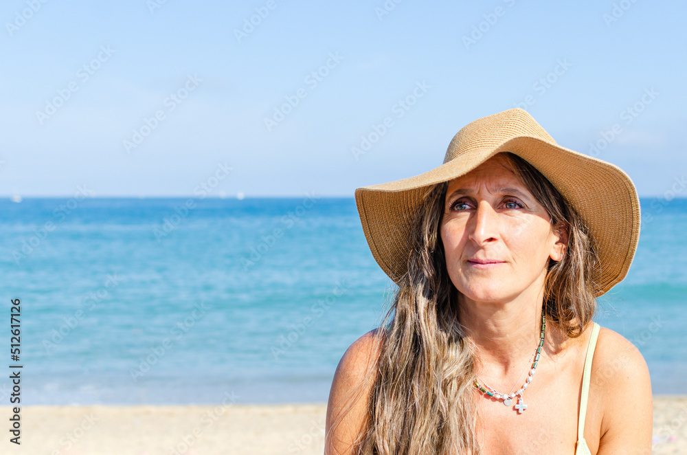 middle-aged woman sitting on the beach. she is wearing a beach hat and a necklace in the shape of a cross. she is looking to the side in concentration, enjoying her free time.