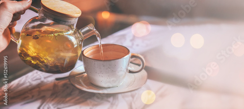 Banner of grey cup with teapot. The hand holding the teapot pours herbal tea into pot on saucer in soft focus on blurred background. Coffee, tea house, bokeh lights. The concept of cozy pastime photo
