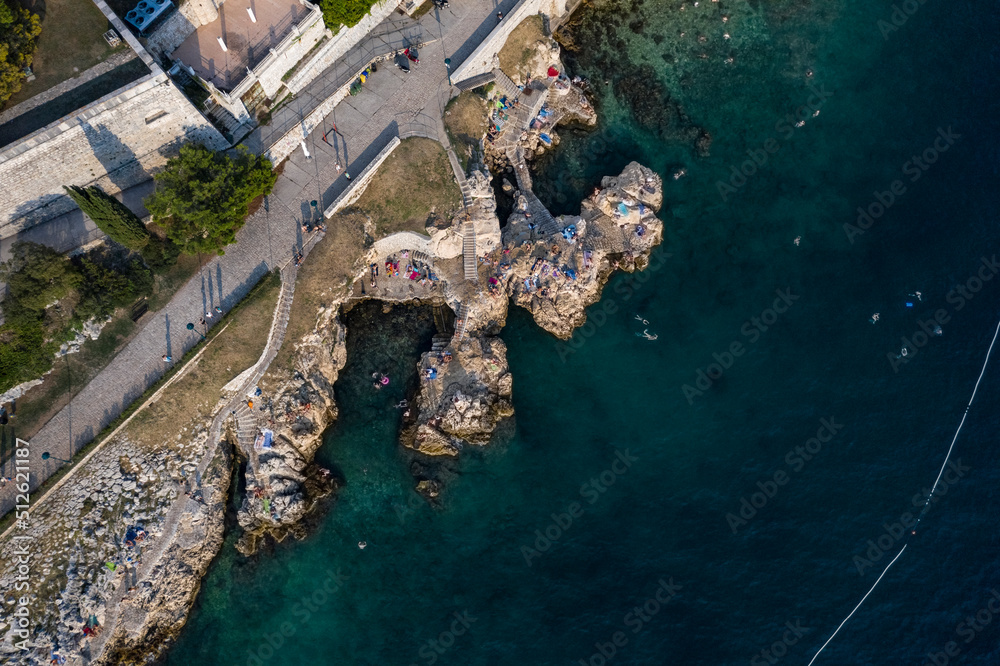 Top aerial view of a rock beach with beautiful blue green water in Rovinj on the Mediterranean sea with road and buildings on the coast