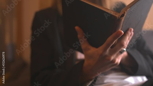 Young man in black suit reading a vintage book on a windowsill. Guy touches aged book to feel the texture of a cover. Dissertation writing on an ancient volume of a rare edition, education process.