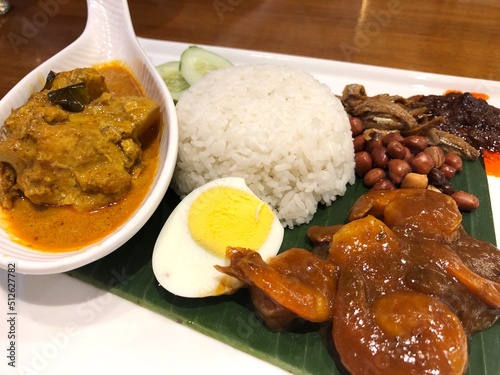 Nasi lemak, a traditional Malay curry paste with fragrant coconut milk tasty rice dish with boiled egg, peanuts, Chili, anchovies, cucumber and prawn sauce. Served on a banana leaf in white plate 