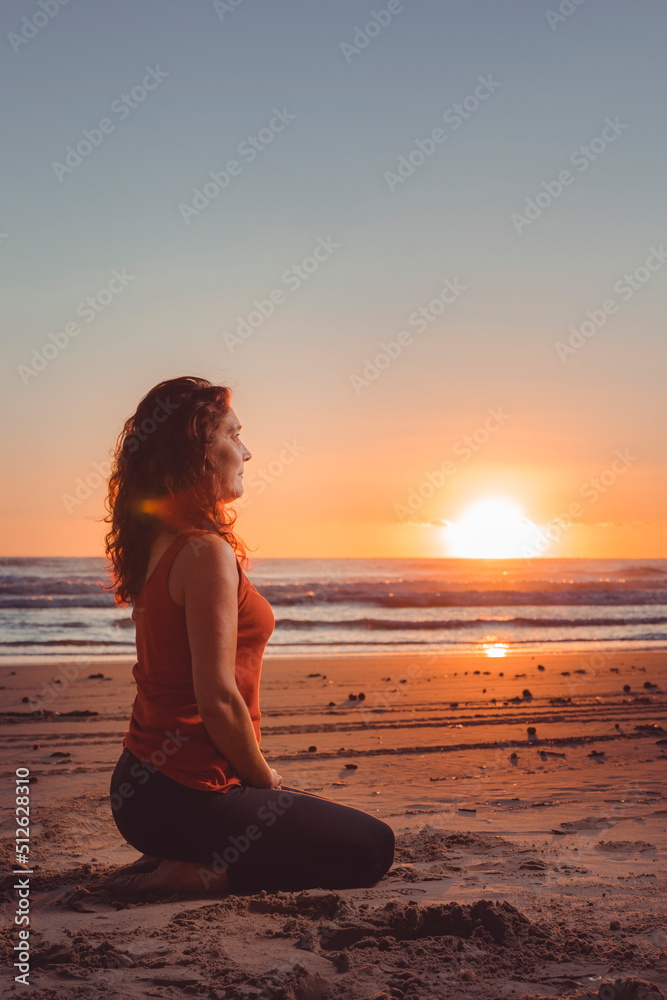 middle-aged woman meditating in front of the sunrise sun on the sand of the beach