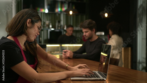 Barista receiving orders on phone browsing online with laptop at cafe. Small business concept of staff using computer at coffee shop