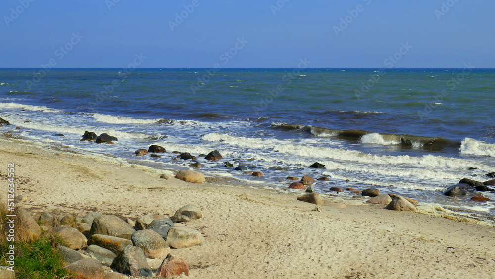 Strand der Ostsee auf der Insel Fehmarn mit Wellen, Sand und großen Kieselsteinen unter blauem Himmel Strand der Ostsee auf der Insel Fehmarn mit Wellen, Sand und großen Kieselsteinen unter blauem Himmel