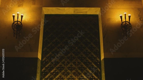 Huge metal medieval castle door in Dark place . Old vintage chandeliers , lamps or lanterns on the wall . Big tall antique door inside church , cathedral , palace or house . Camera moving on door