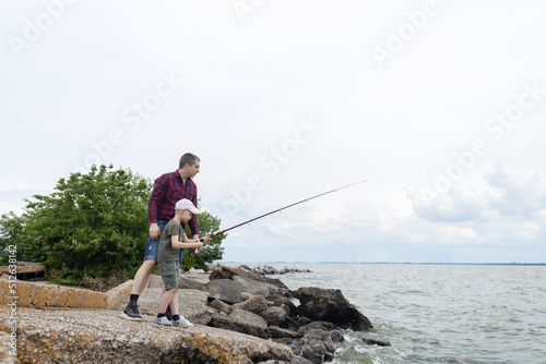 Father and son fishing. Dad shows his son how to hold the spinning and spin the reel. Fishing training on a pond or river. Caring parent concept.