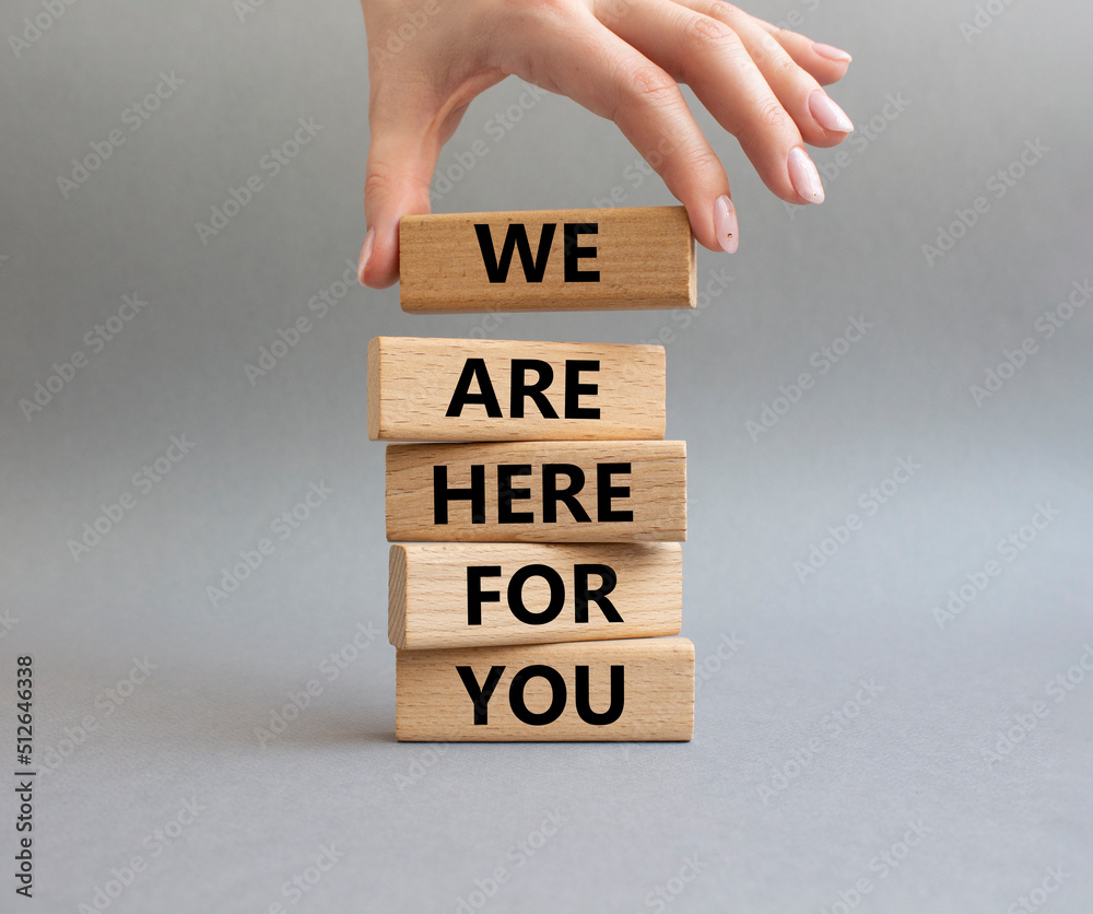Help symbol. Wooden blocks with words 'We are here for you'. Beautiful grey background. Businessman hand. Business and 'We are here for you' concept. Copy space. Help symbol. Wooden blocks with words 'We are here for you'. Beautiful grey background. Businessman hand. Business and 'We are here for you' concept. Copy space.