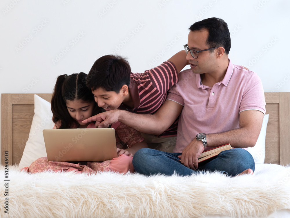 Fototapeta premium Indian father's hand pointing to the laptop for two teenage children ,son and daughter, sitting on the bed together at home. Family using laptop for movie, homework, online learning.