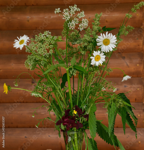 A bouquet of flowering garden weeds - daisies, dandelions, snyt and grasses in a transparent glass vase against a wooden wall made of logs photo