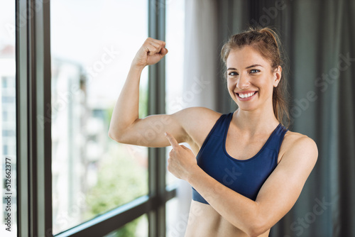 Happy young woman flexing her biceps at home and looking at camera Free Download Happy young woman flexing her biceps at home and looking at camera - GFXLoad