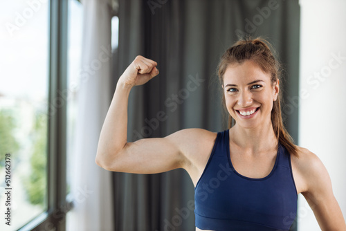 Happy young woman flexing her biceps at home and looking at camera Free Download Happy young woman flexing her biceps at home and looking at camera - GFXLoad