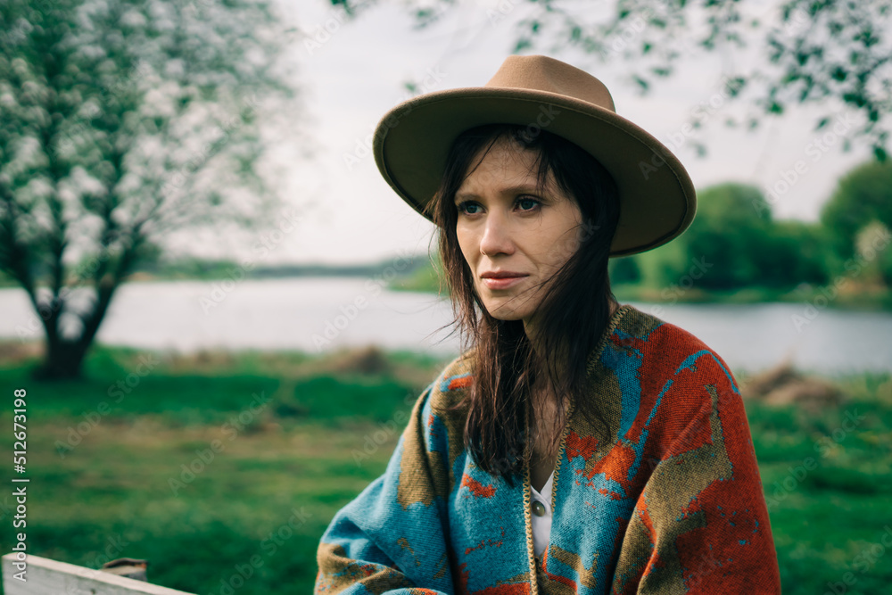 Woman in a hat and poncho at cloudy autumn day stands on countryside with river.
