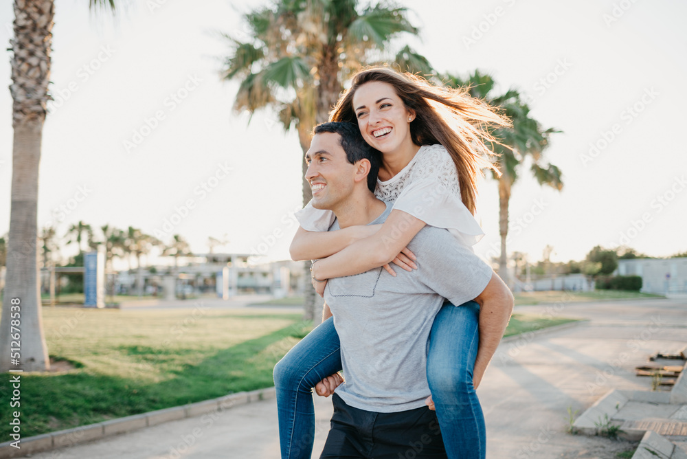A smiling brunette girl is riding her Hispanic boyfriend between palms in Spain
