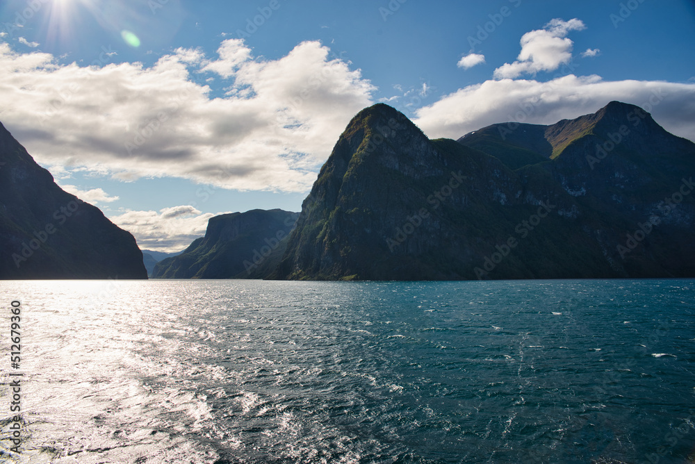 View from a ferry in Sognefjord, Norway with high mountains rising directly out of the water on a beautiful and sunny day with clouds above the mountains. Summer feelings, vacation time.  