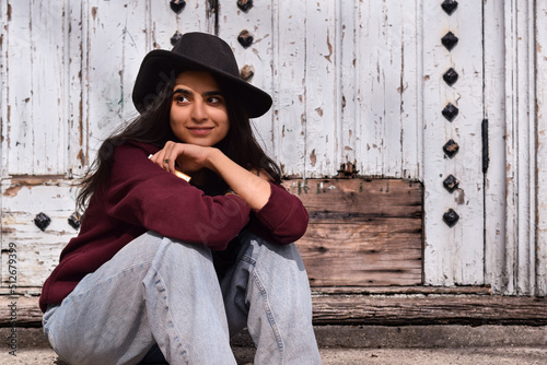 Portrait of young stylish girl in fedora hat sitting on steps of old building smilimg photo