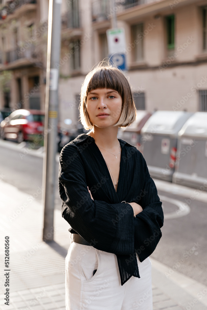 Outside portrait of stylish pretty European girl with straight short hairstyle wearing black blouse and white pants posing at camera in sunlight on summer street