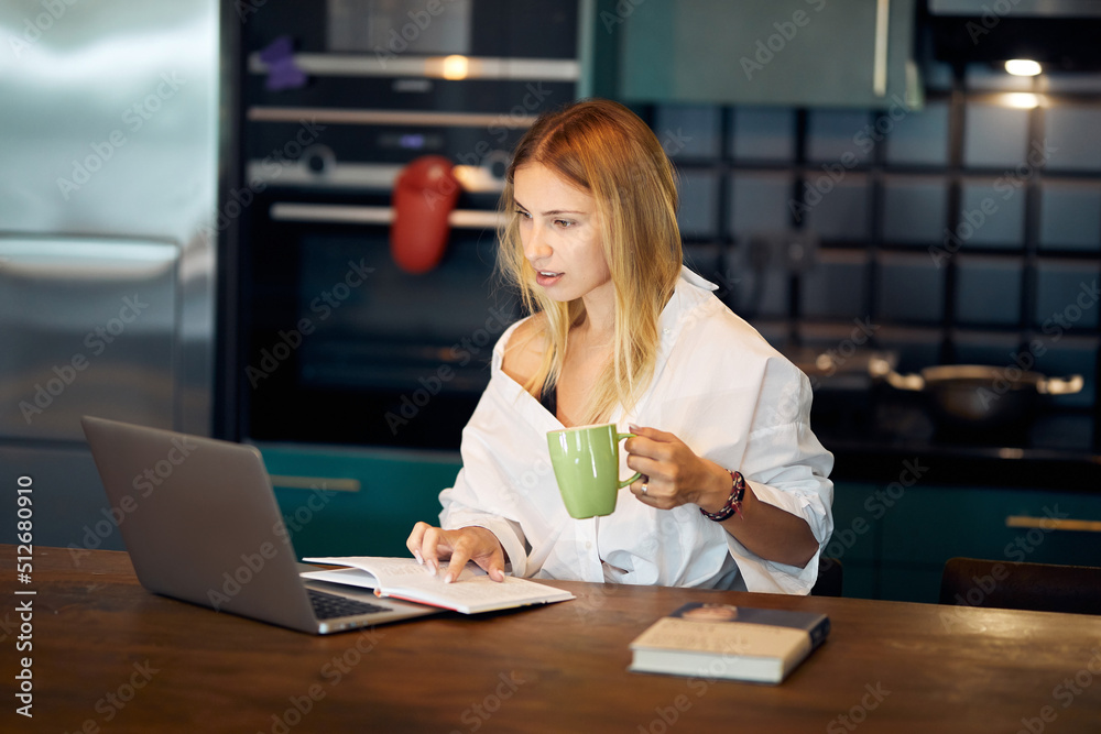 Young woman working at home kitchen looks at notebook with a cup of coffee in a hand Young woman working at home kitchen looks at notebook with a cup of coffee in a hand