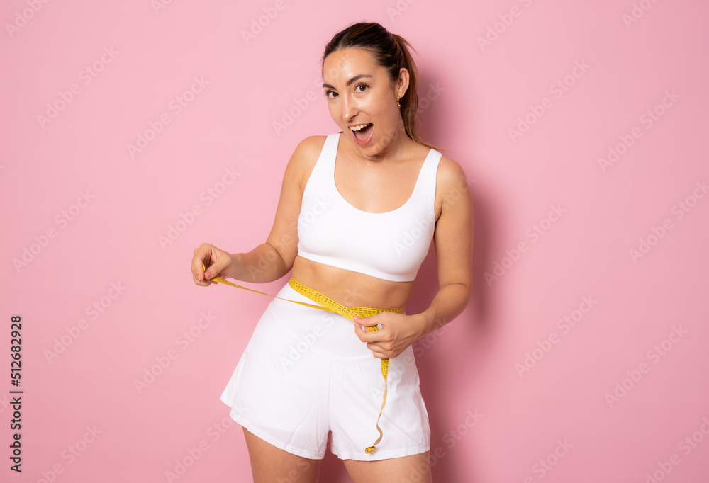 Portrait of a satisfied brunette fitness woman holding measuring tape around her waist isolated over pink background