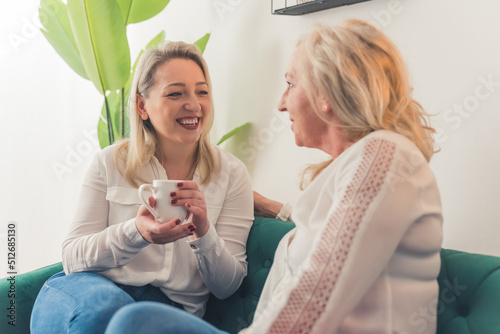 Cheerful blonde beautiful woman in her 30s holding a white mug, chatting with her elegant blonde-haired middle-aged female friend. Indoor shot. High quality photo
