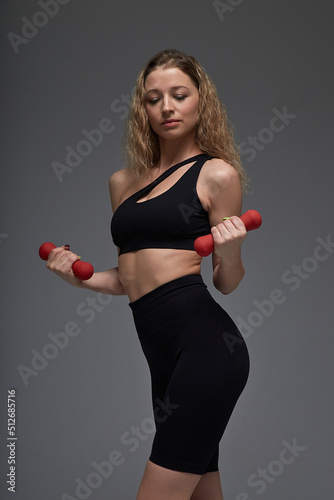 young fit sporty attractive blond caucasian female in black sportswear holding red dumbbells in hands at gray background. sport, fitness training, healthy lifestyle, workout © Artem Zatsepilin