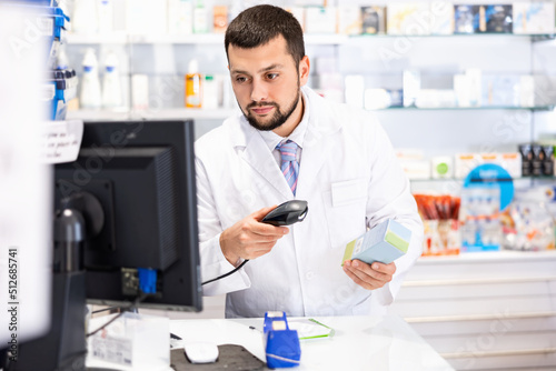 Male pharmacist in lab coat standing at counter in drugstore and using barcode scanner to sale drug package.