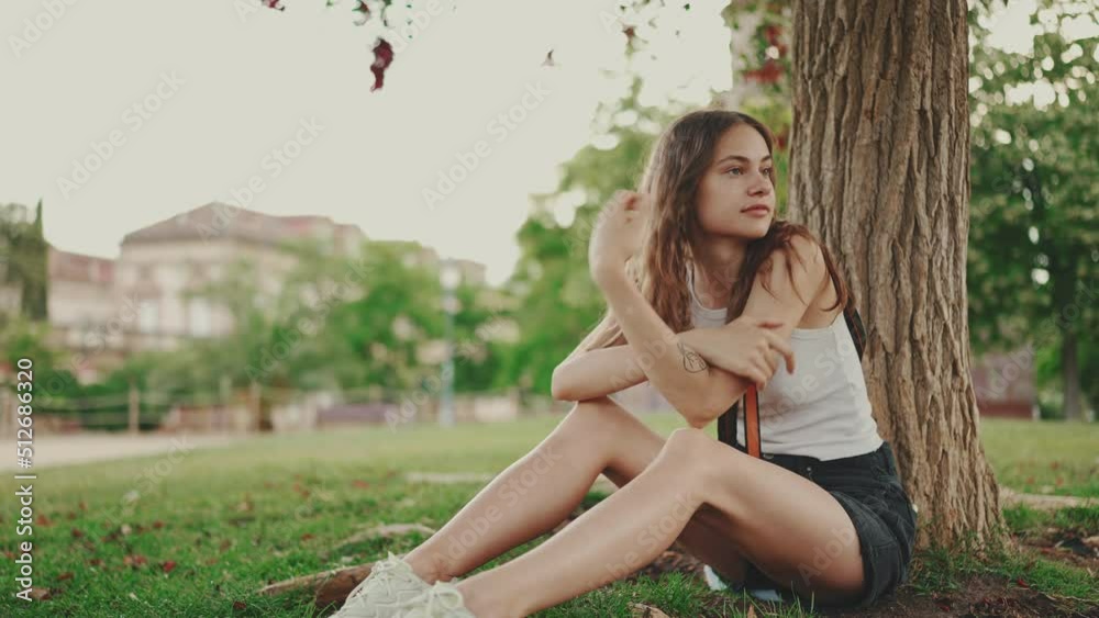 Girl is resting in the park. Beautiful girl looks around sitting on the lawn under tree in the park Girl is resting in the park. Beautiful girl looks around sitting on the lawn under tree in the park