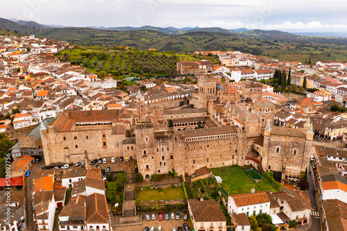 Gothic-Mudejar style building of Royal Monastery of Saint Mary in Spanish town of Guadalupe, located in green valley of province of Caceres overlooking brownish roofs of houses, as seen from drone.. photo