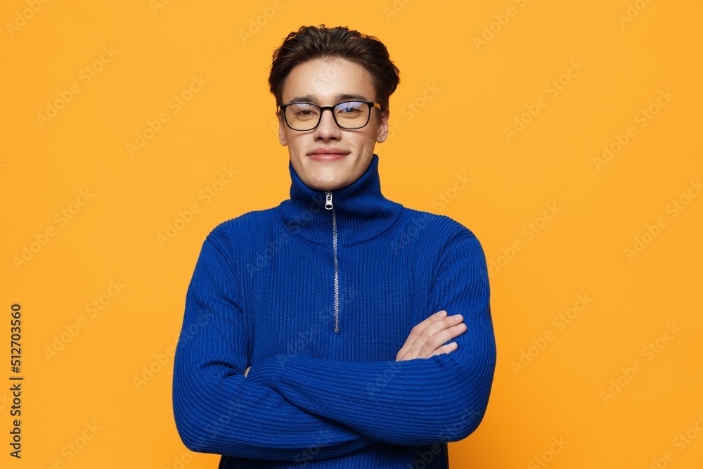 an attractive young man stands on an orange background in a blue zip-up jacket and black eyeglasses and looks into the camera with a pleasant smile holding his arms crossed on his chest an attractive young man stands on an orange background in a blue zip-up jacket and black eyeglasses and looks into the camera with a pleasant smile holding his arms crossed on his chest