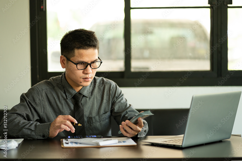 An Asian male accountant who works from home is analyzing market data and has a calculator to calculate his company's finances. In a private office at home, New Normal concept. An Asian male accountant who works from home is analyzing market data and has a calculator to calculate his company's finances. In a private office at home, New Normal concept.