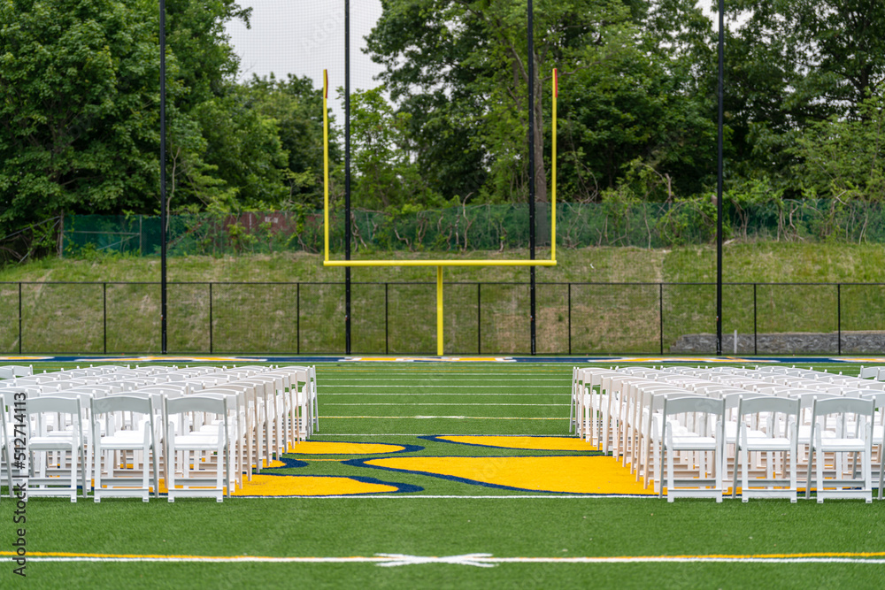 Fototapeta premium White chairs set-up in rows on a green synthetic turf athletic field for a high school graduation ceremony. 