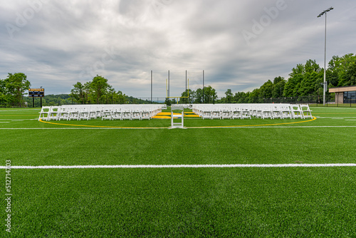 White folding chairs set-up in rows, with single chair at center, on a green synthetic turf athletic field for a high school graduation ceremony.  © Thomas