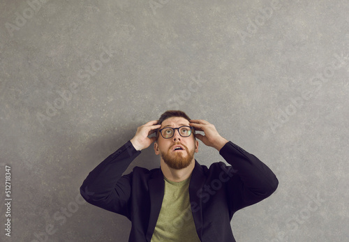 Young man afraid of something. Shocked scared anxious guy in hipster glasses looking up at terrible threat and danger sitting against grey concrete studio wall. Fear, fright, problem, trouble concept photo