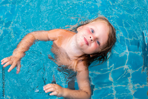 A little girl with blue eyes is bathing in the blue water of the pool. Summer children's games. Happy child