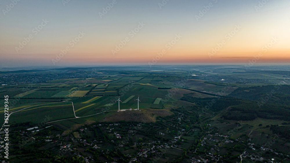 aerial shot of two wind turbines on a hill over a village during sunrise