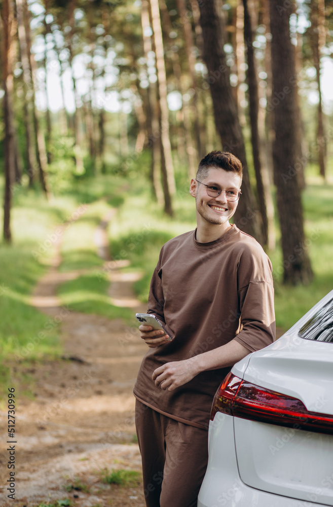 A young guy with a beard talking on the phone near his electric car. In his hands he has a fashionable smartphone. He has a good mood.