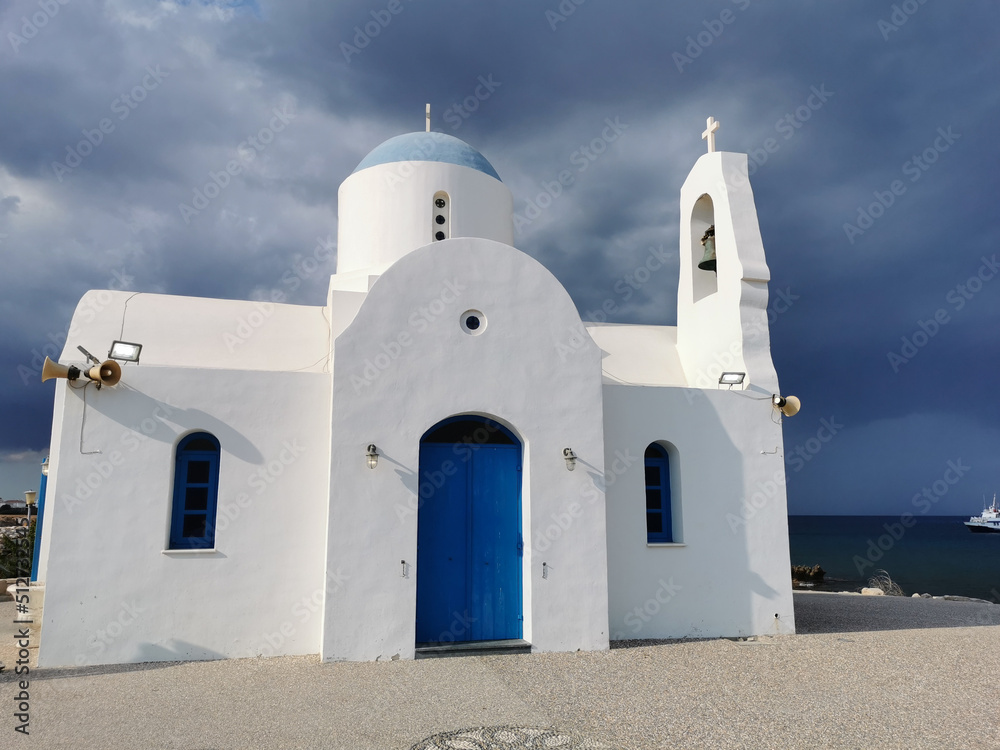 The Church of St. Nicholas the Wonderworker is white with a blue door against the backdrop of the Mediterranean Sea and the dramatic sky.