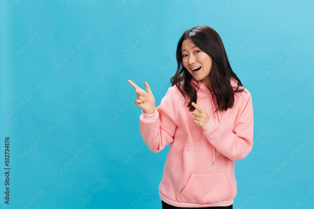 Overjoyed cute Asian student young lady in pink hoodie sweatshirt point fingers aside posing isolated on over blue studio background. The best offer for ad. People Emotions for Everyday concept