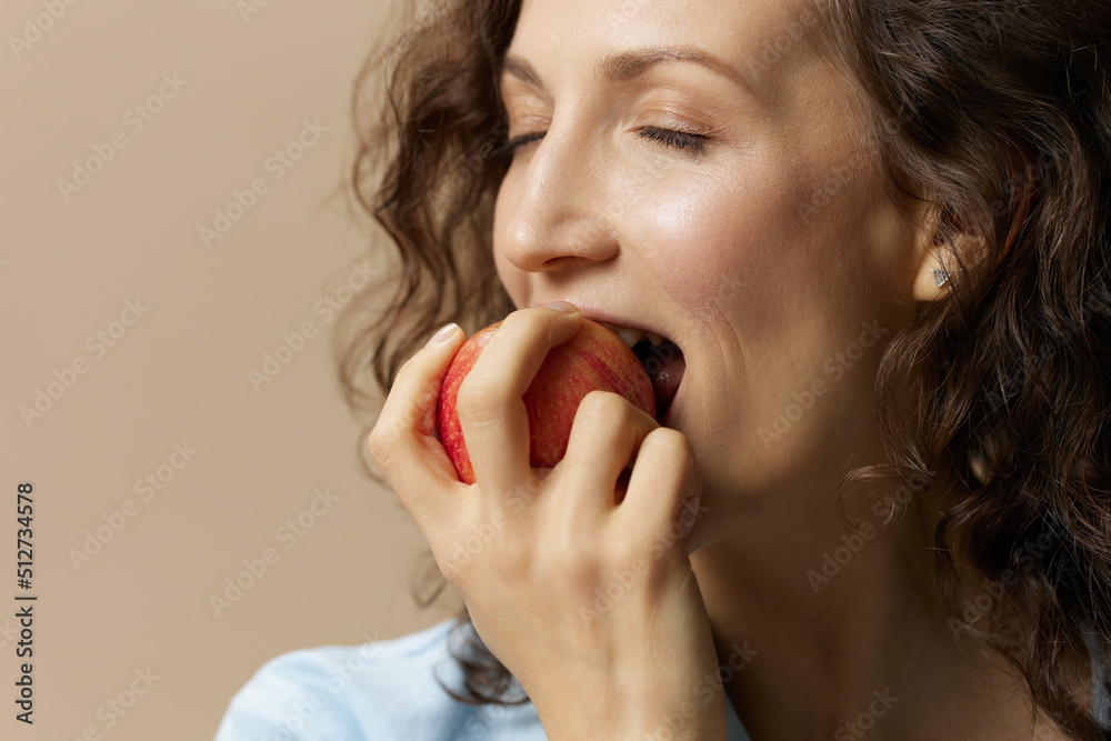 Cute happy curly beautiful female in jeans casual shirt bites apple enjoy sweet fruit posing isolated on over beige pastel background. Healthy food. Natural eco-friendly products concept. Copy space Cute happy curly beautiful female in jeans casual shirt bites apple enjoy sweet fruit posing isolated on over beige pastel background. Healthy food. Natural eco-friendly products concept. Copy space